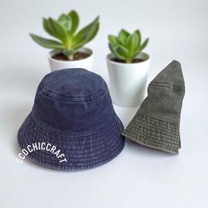 May include: A navy blue bucket hat with a washed finish and the white text ECOCHICCRAFT printed on the front, positioned next to a folded olive green bucket hat. Both hats feature a circular crown and a downward-sloping brim with stitched detailing.