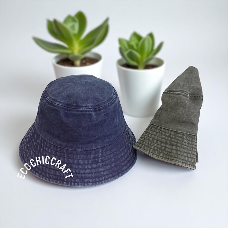 May include: A navy blue bucket hat with a washed finish and the white text ECOCHICCRAFT printed on the front, positioned next to a folded olive green bucket hat. Both hats feature a circular crown and a downward-sloping brim with stitched detailing.