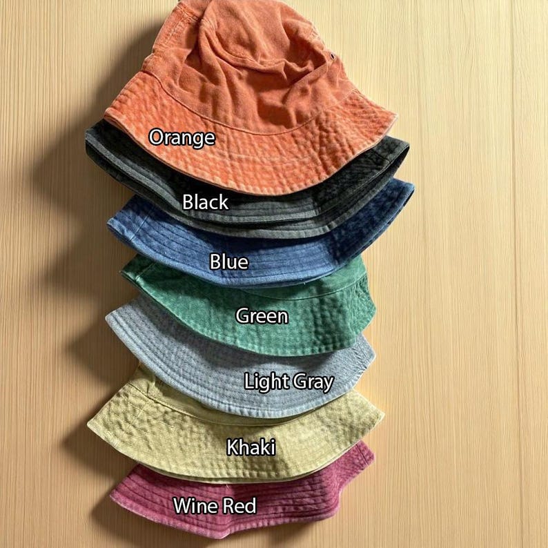 May include: A stack of seven bucket hats in various colors, labeled with text: Orange, Black, Blue, Green, Light Gray, Khaki, and Wine Red. Each hat features a wide, downward-sloping brim and a rounded crown, showcasing a consistent design across the collection.