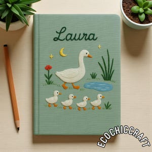 Puede incluir: Un cuaderno de color verde salvia con bordados de una oca blanca, cuatro patitos, un estanque azul, una flor roja, una luna creciente amarilla y estrellas amarillas. El nombre Laura est&aacute; bordado en cursiva verde oscuro en la portada.