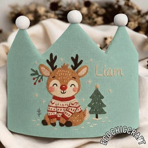 May include: A teal crown with three white pom-poms. It features an embroidered reindeer in a sweater, a Christmas tree, and the name "Liam". The base of the crown has the text "ECOCHICCRAFT".