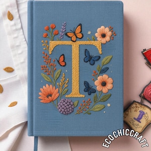 Peut inclure: Un journal bleu avec un motif brodé représentant la lettre "T", des papillons et des fleurs orange, jaunes et bleues. Le journal a un signet ruban bleu assorti.