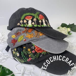 May include: Three embroidered baseball caps stacked on top of each other. The top cap is black with a mushroom design. The middle cap is gray with an autumn leaves design. The bottom cap is black with a floral design and the text "ECOCHIC CRAFT".