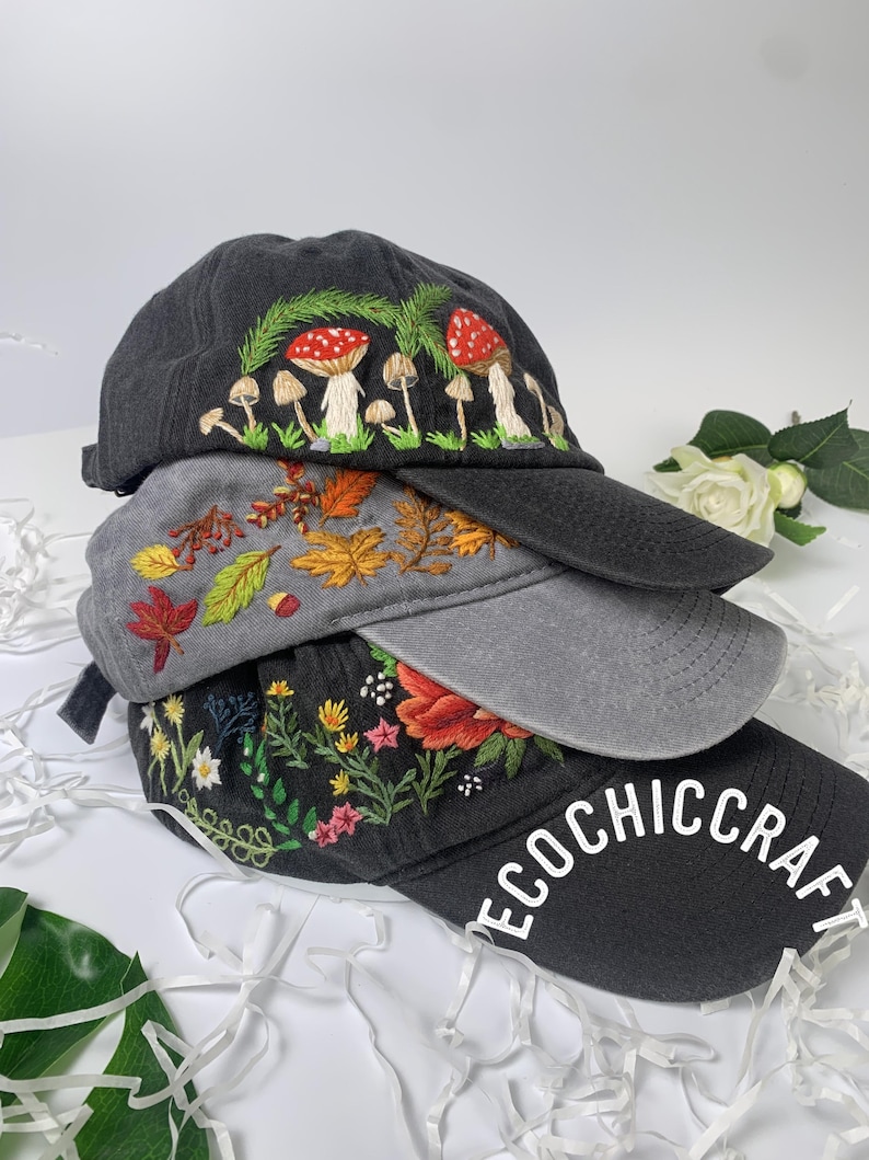 May include: Three embroidered baseball caps stacked on top of each other. The top cap is black with a mushroom design. The middle cap is gray with an autumn leaves design. The bottom cap is black with a floral design and the text "ECOCHIC CRAFT".