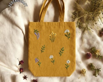 May include: Mustard yellow tote bag with embroidered floral designs. The bag features embroidered flowers and leaves in white, yellow, and purple. The bag has two handles. The text "ECOCHICCRAFT" is visible.