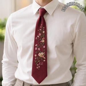 Può includere: Una cravatta bordeaux scuro con un intricato ricamo floreale nei toni del rosa, giallo e verde. Il design presenta un motivo a vite rampicante con piccoli fiori e le iniziali E.L. ricamate vicino alla parte inferiore. Il testo ECOCHICCRAFT è visibile.