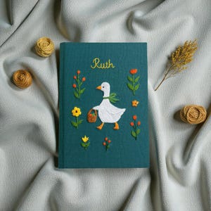 Puede incluir: Un diario cubierto de tela verde azulado oscuro con detalles bordados a mano. La portada muestra una oca blanca con una bufanda verde y una cesta marr&oacute;n, rodeada de flores amarillas y naranjas. El nombre Ruth est&aacute; bordado en amarillo.