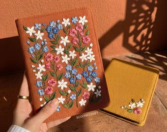 May include: A rectangular notebook cover in burnt orange fabric featuring dense floral embroidery in white, blue, and pink. A second mustard yellow notebook cover sits nearby with a small floral embroidery cluster. The text ECOCHICCRAFT is visible.