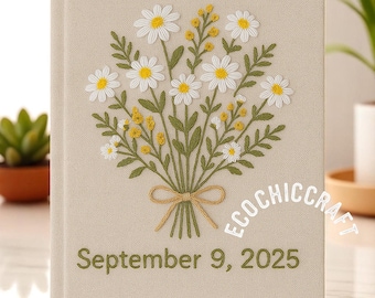 May include: A beige linen journal with a hand-stitched design of a bouquet of white daisies and green leaves. The journal has the date "September 9, 2023" embroidered on the front.