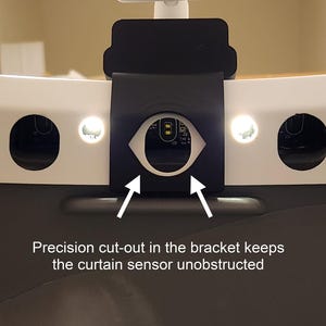 May include: Close-up of a black bracket with a precision cut-out, designed to keep the curtain sensor unobstructed. The bracket is mounted on a white surface with two circular openings and two small lights. Text on the image reads: "Precision cut-out in the bracket keeps the curtain sensor unobstructed."