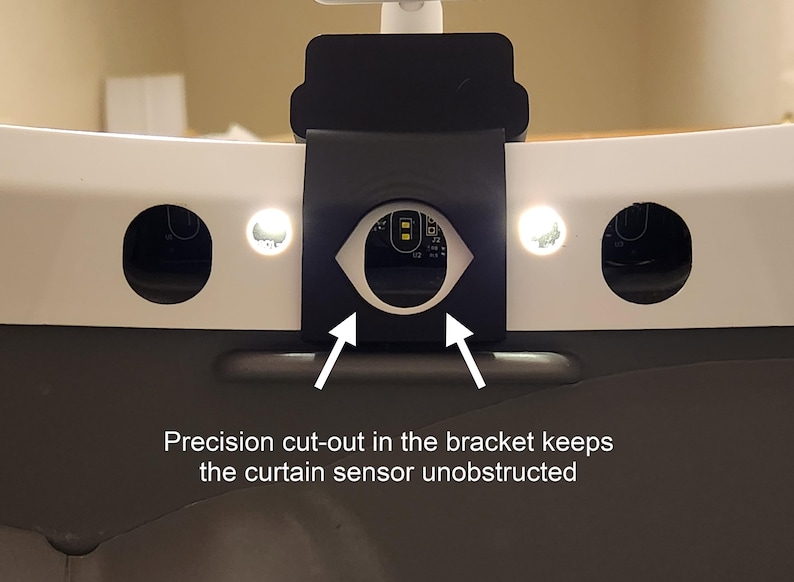 May include: Close-up of a black bracket with a precision cut-out, designed to keep the curtain sensor unobstructed. The bracket is mounted on a white surface with two circular openings and two small lights. Text on the image reads: "Precision cut-out in the bracket keeps the curtain sensor unobstructed."