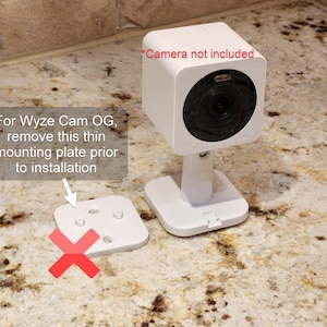 May include: A white Wyze Cam OG security camera with a square design, mounted on a stand. The image includes text that says "Camera not included" and instructions to remove a mounting plate before installation.