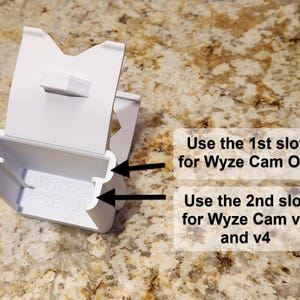 May include: A white plastic Wyze camera mount with two slots. Text on the image reads: "Use the 1st slot for Wyze Cam OG" and "Use the 2nd slot for Wyze Cam v3 and v4". The mount is designed to hold Wyze cameras.