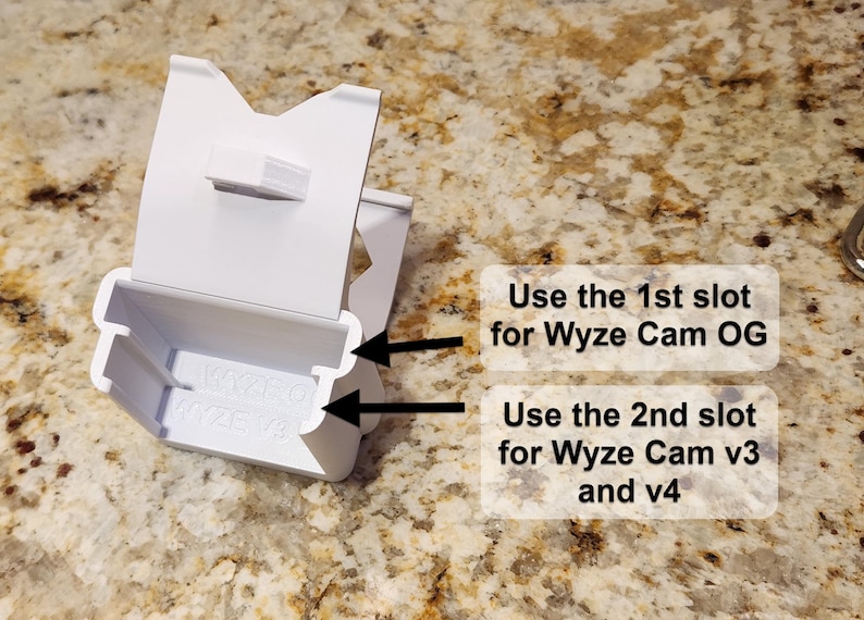 May include: A white plastic Wyze camera mount with two slots. Text on the image reads: "Use the 1st slot for Wyze Cam OG" and "Use the 2nd slot for Wyze Cam v3 and v4". The mount is designed to hold Wyze cameras.