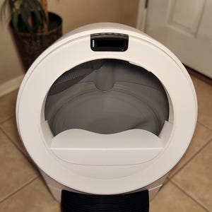 Op de afbeelding: Een witte, bolvormige automatische kattenbak met een grote ronde opening, een grijze binnenbak, een zwart digitaal displaypaneel aan de bovenkant en een zwarte geribbelde opstapmat aan de onderkant van de unit voor in- en uitgang.