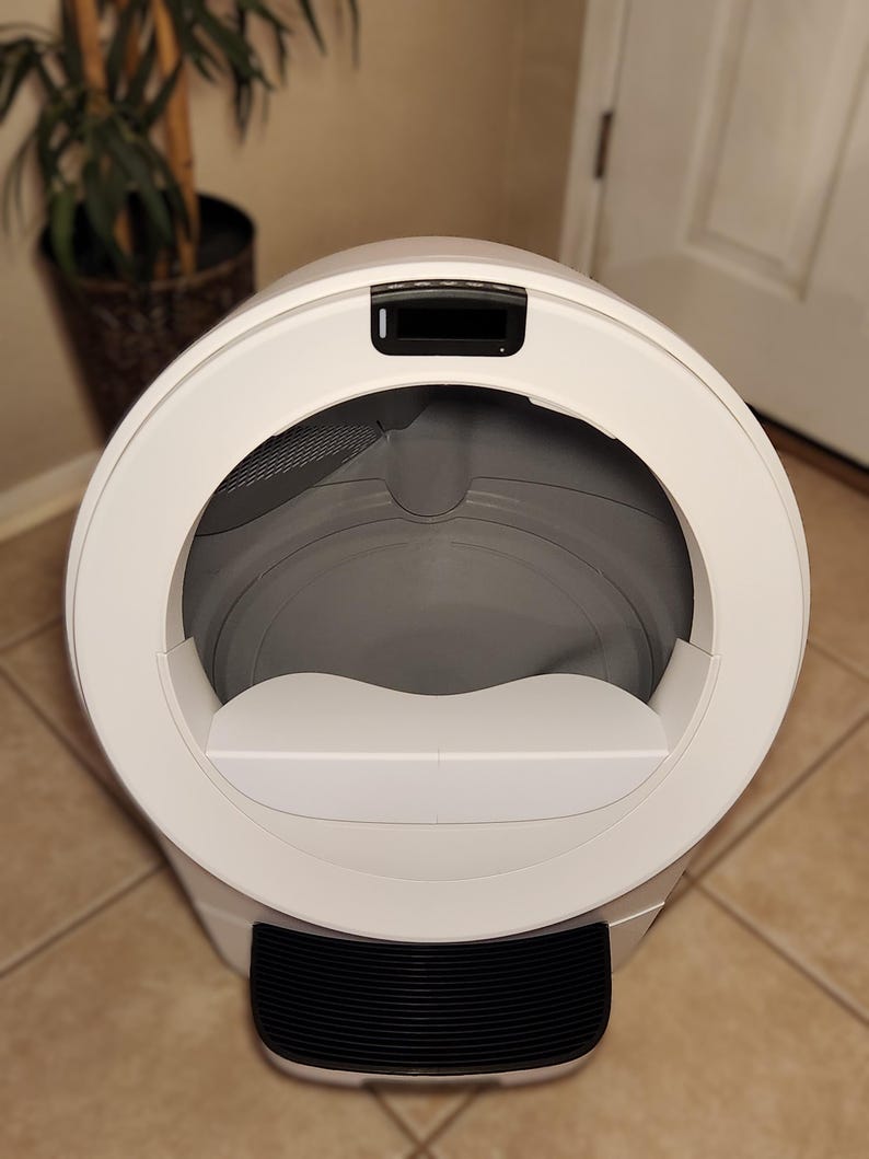 Op de afbeelding: Een witte, bolvormige automatische kattenbak met een grote ronde opening, een grijze binnenbak, een zwart digitaal displaypaneel aan de bovenkant en een zwarte geribbelde opstapmat aan de onderkant van de unit voor in- en uitgang.