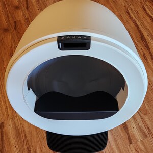 Op de afbeelding: Een geautomatiseerde, eivormige kattenbak met een gladde witte buitenkant. De voorzijde is voorzien van een grote ronde opening, een zwarte binnenbak, een zwart bedieningspaneel met vijf knoppen en een zwarte geribbelde opstap.