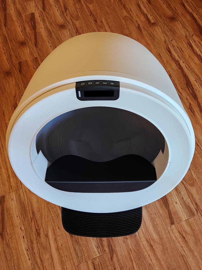 Op de afbeelding: Een geautomatiseerde, eivormige kattenbak met een gladde witte buitenkant. De voorzijde is voorzien van een grote ronde opening, een zwarte binnenbak, een zwart bedieningspaneel met vijf knoppen en een zwarte geribbelde opstap.