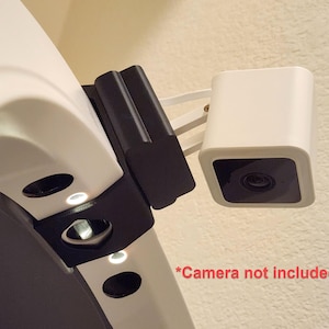 May include: A white security camera with a black lens is mounted on a wall. The camera is attached to a black bracket and a white arm. The text "*Camera not included" is visible in red.