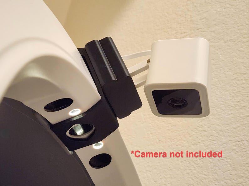 May include: A white security camera with a black lens is mounted on a wall. The camera is attached to a black bracket and a white arm. The text "*Camera not included" is visible in red.