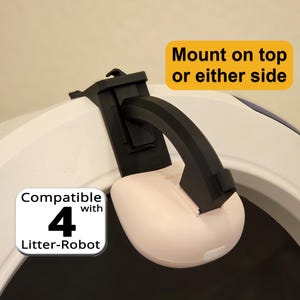 May include: A black plastic mounting bracket attached to a white curved surface, featuring a light pink rounded housing. Text on the image reads: Mount on top or either side and Compatible with 4 Litter-Robot.