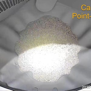 May include: An overhead view inside a washing machine drum filled with soapy water and suds. The text "Camera Point-of-View" is in the upper right corner, with the disclaimer "*Wyze cameras sold separately" below it. The date and time are displayed at the bottom.