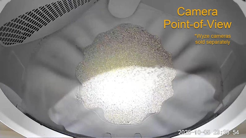 May include: An overhead view inside a washing machine drum filled with soapy water and suds. The text "Camera Point-of-View" is in the upper right corner, with the disclaimer "*Wyze cameras sold separately" below it. The date and time are displayed at the bottom.