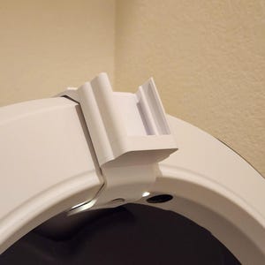 May include: A white, 3D-printed object, possibly a bracket or clip, attached to a curved white surface. The object has a rectangular shape with a series of vertical lines and a small opening. The background is a neutral beige color.