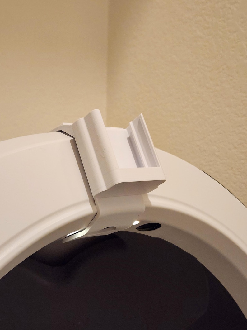 May include: A white, 3D-printed object, possibly a bracket or clip, attached to a curved white surface. The object has a rectangular shape with a series of vertical lines and a small opening. The background is a neutral beige color.
