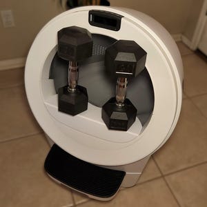 Op de afbeelding: Een wit cirkelvormig fitnessapparaat met een zwart digitaal display aan de bovenkant en een zwarte geribbelde trede aan de basis. Twee zwarte zeshoekige halters met metallic zilveren handgrepen zijn in de centrale opening geplaatst.