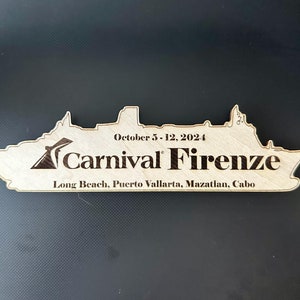 May include: A wooden cutout of a cruise ship with the text "October 5-12, 2024 Carnival Firenze Long Beach, Puerto Vallarta, Mazatlan, Cabo" engraved on it.