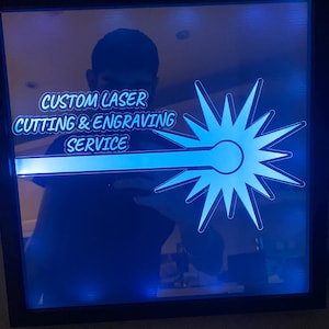 Könnte beinhalten: Ein gerahmtes beleuchtetes Schild mit blauem Hintergrund und einem weißen Sternenausbruch-Design. Das Schild lautet "Custom Laser Cutting & Engraving Service".