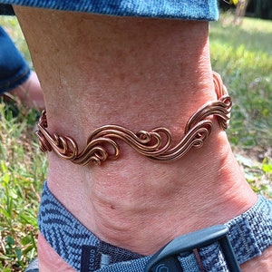 May include: A copper-colored anklet with a flowing, wave-like design. The anklet is worn on an ankle, paired with a blue and black sandal. The background features green grass and sunlight.
