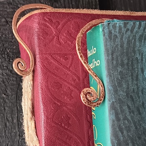 May include: A handmade copper bookmark with a hammered texture and a spiral design, placed on the corner of a red leather-bound book. The book has embossed patterns and a teal-colored page edge.