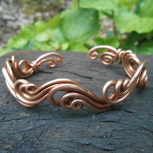 May include: A copper wire bracelet with a wavy, swirling design. The bracelet is made of multiple strands of wire that are twisted and woven together to create a unique and intricate pattern.