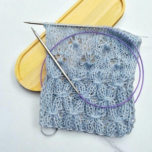 Peut inclure: Gros plan sur un tissu tricoté bleu clair avec un motif texturé, avec des aiguilles à tricoter argentées et une aiguille circulaire violette. Le tissu est partiellement terminé, avec des mailles encore sur les aiguilles. Un plateau en bois est en arrière-plan.