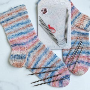 May include: A pair of hand-knitted socks in pastel colors of blue, pink, and peach, with a set of circular knitting needles and a stitch marker. The socks are made with a striped pattern.
