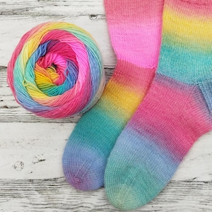 Könnte beinhalten: Ein Paar handgestrickte Socken mit einem Regenbogen-Farbverlauf, der von Rosa über Gelb, Grün und Blau verläuft. Eine passende Garnkugel liegt neben den Socken und zeigt ebenfalls ein lebendiges Farbspektrum.
