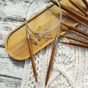 May include: A set of bamboo knitting needles with a cable and stitch markers on a wooden tray. The needles are arranged in a fan shape and are ready for use.