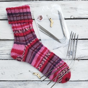 May include: A hand-knitted sock in shades of pink, red, and purple, with a folded cuff. The sock is accompanied by a set of knitting needles and small wooden sock-shaped stitch markers. A clear plastic bag contains more needles.