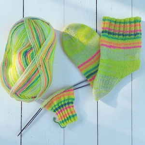 Puede incluir: Un ovillo de lana multicolor y calcetines parcialmente tejidos en tonos verde neón, rosa, amarillo y azul. La lana es una vibrante mezcla de colores, y los calcetines están en varias etapas de finalización, con agujas de tejer visibles.