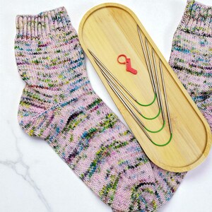 Puede incluir: Un par de calcetines tejidos a mano en un patr&oacute;n moteado rosa, verde y azul. Una bandeja de madera contiene varias agujas de tejer plateadas con cables verdes y un marcador de punto rojo. Los calcetines se muestran sobre una superficie blanca.