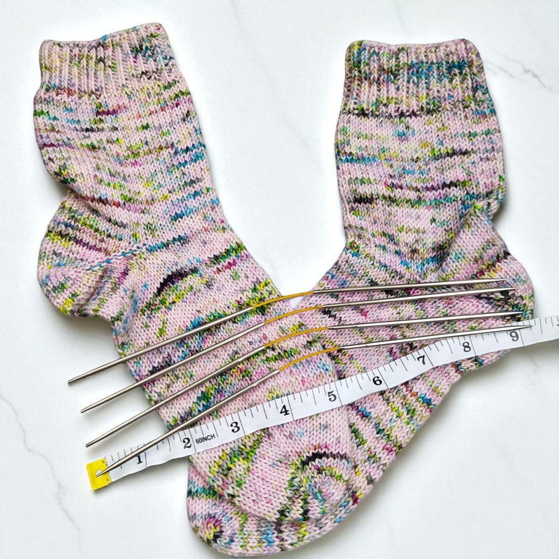 Puede incluir: Un par de calcetines tejidos a mano con un patr&oacute;n moteado rosa, verde y azul. Los calcetines se miden con una cinta m&eacute;trica, mostrando una longitud de aproximadamente 23 cm. Las agujas de tejer se colocan junto a los calcetines.