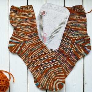Puede incluir: Calcetines tejidos a mano en tonos marr&oacute;n, naranja y azul, con un patr&oacute;n de rayas. Tambi&eacute;n se ven agujas de tejer y una madeja de lana naranja, lo que sugiere un tema de artesan&iacute;a.