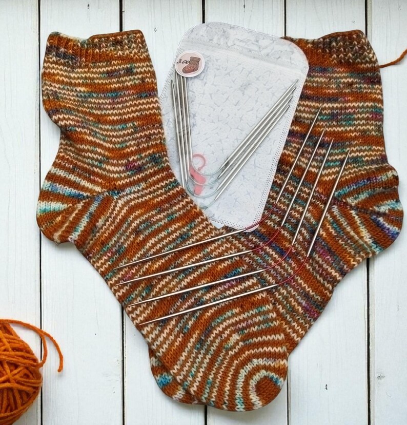 Puede incluir: Calcetines tejidos a mano en tonos marr&oacute;n, naranja y azul, con un patr&oacute;n de rayas. Tambi&eacute;n se ven agujas de tejer y una madeja de lana naranja, lo que sugiere un tema de artesan&iacute;a.