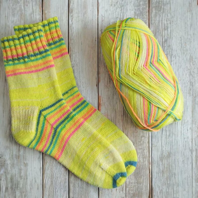 Peut inclure: Une paire de chaussettes jaunes avec des rayures horizontales roses, oranges, bleues et vertes. Une pelote de laine assortie est &agrave; c&ocirc;t&eacute; des chaussettes. Le fil et les chaussettes sont sur une surface en bois clair.