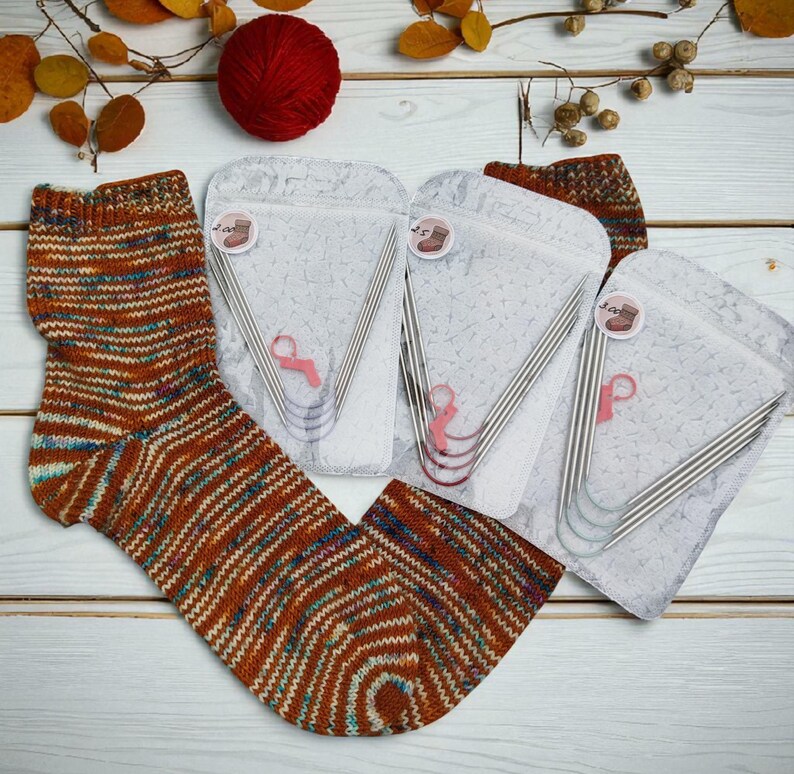 Puede incluir: Calcetines tejidos a mano con rayas marrones, naranjas y azules. Se ven varios juegos de agujas de tejer en fundas transparentes. Una madeja de lana roja y follaje decorativo completan la escena sobre una superficie de madera blanca.