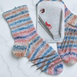 May include: A pair of hand-knitted socks with a colorful striped pattern in shades of blue, pink, and orange. Knitting needles and a stitch marker are included, along with a small package with a sock icon and the number 2.5.