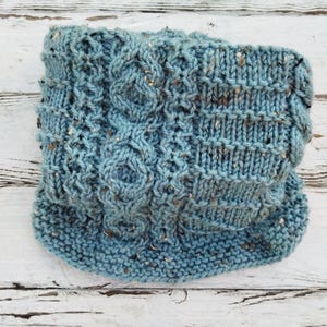 May include: A blue knitted neck warmer with a cable knit pattern and flecks of white throughout.