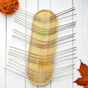 Puede incluir: Un conjunto de agujas de tejer circulares con cables flexibles de varios colores, dispuestas sobre una bandeja de madera. Una madeja de lana naranja y una hoja de oto&ntilde;o tambi&eacute;n est&aacute;n presentes. Las agujas son de metal.